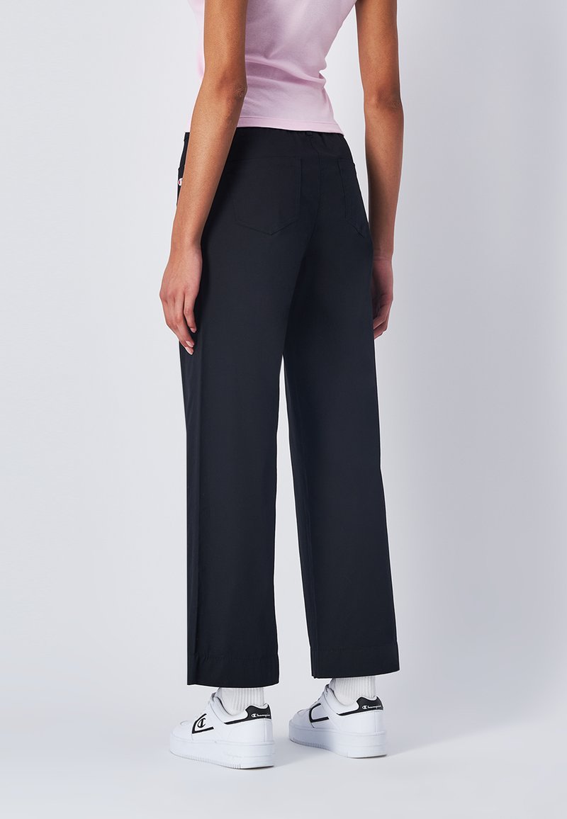 Pantaloni neri a gamba larga con vita alta, dotati di due tasche posteriori, texture liscia e orlo pulito, abbinati a sneaker bianche e calzini.