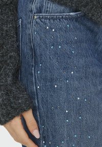 Blugi din denim cu o croială ajustată, împodobiți cu strasuri albastre, argintii și negre, dispersate pe față, lângă un pulover texturat de culoare închisă.