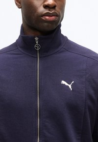 Posnetek v bližini osebe, ki nosi mornarsko modro Puma jakno z zadrgo in belim Puma logotipom na levi strani prsnega koša.