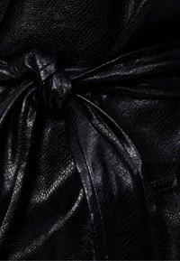 Czarna tkanina z sztucznej skóry o teksturowanym wykończeniu, z węzełkowym detalem tworzącym fałdy i draperie. Gładki i błyszczący wygląd.