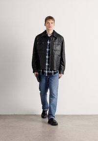 Black leather jacket with two front pockets, worn over a blue plaid shirt. Paired with light blue jeans and black shoes. Model holds a black device.