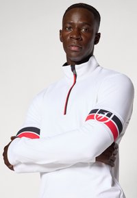 Top sportiv alb cu fermoar și mâneci lungi. Prezintă accente roșii și negre pe brațe, un guler cu fermoar și un material texturat și neted.