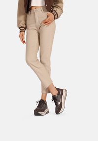 Pantaloni beige, con trama, aderenti abbinati a sneaker sportive scure caratterizzate da un mix di materiali e un logo sottile. Una giacca bomber marrone esalta il look.