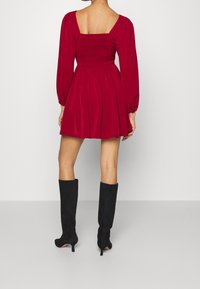 Rotes Mini-Kleid mit gerafftem Oberteil und langen Puffärmeln; quadratischer Rückenausschnitt. Kombiniert mit knielangen schwarzen Stiefeln aus Wildleder mit Absatz.