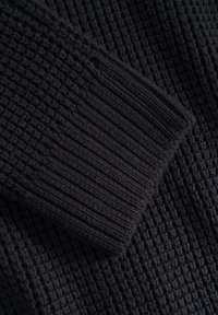 Czarna dzianina z teksturowanym, prążkowanym wzorem. Zbliżenie na mankiet, ukazujące grubą przędzę i gęsty wzór.