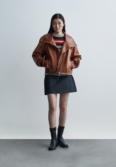 Brązowa skórzana kurtka motocyklowa z szerokim kołnierzem, zapinana na suwak, noszona na swetrze z dzianiny i ciemnej jeansowej mini spódnicy, w połączeniu z czarnymi botkami za kostkę.