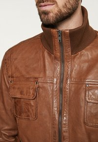 Brown leather jacket featuring a ribbed collar, front zip closure, and two chest pockets with snap buttons. The texture is smooth and glossy.