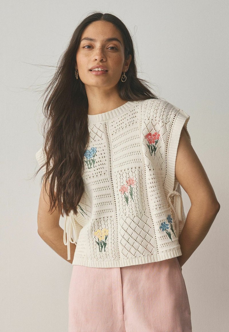 Femme aux longs cheveux foncés portant un haut en maille blanc sans manches avec des motifs floraux colorés et un pantalon rose clair, debout devant un fond uni.