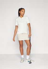 White short-sleeve polo shirt with cable knit texture, beige golf skirt, and light gray sneakers with teal accents. Golf club held in left hand.