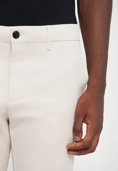 Pantaloni bej deschis cu o textură netedă, având o închidere cu un nasture negru și buzunare laterale, arătați aproape de o mână pe talie.