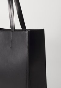 Sac à main en cuir noir avec une texture lisse, une forme structurée et des poignées plates doubles. Présente un design minimaliste et des détails de couture.