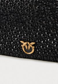 Clutch negru adornat cu pietre negre grupate și un mic emblemă aurie care înfățișează două păsări față în față pe un inel.
