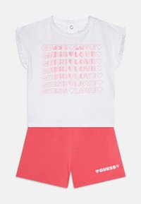 Weißes kurzärmliges Oberteil mit pinkem Glitzertext "GUESS ♥ LOVE", der wiederholt erscheint. Korallfarbene Shorts mit "GUESS ♥" in Weiß auf dem Bein gedruckt. Aus weichem Baumwollstoff.