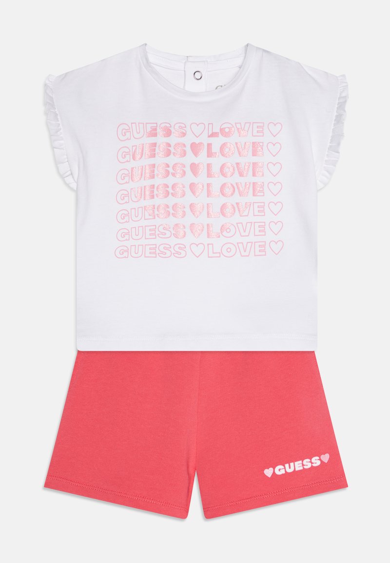 Weißes kurzärmliges Oberteil mit pinkem Glitzertext "GUESS ♥ LOVE", der wiederholt erscheint. Korallfarbene Shorts mit "GUESS ♥" in Weiß auf dem Bein gedruckt. Aus weichem Baumwollstoff.