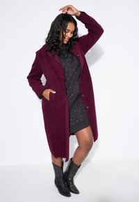 Cappotto in lana bordeaux con design su misura, rever a punta e chiusure a bottoni. Indossato sopra un abito nero con pois bianchi e stivali neri.