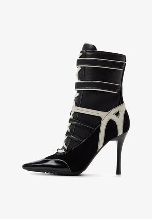 Black high-heeled ankle boot with pointed toe, lace-up front, white and black straps, and white geometric detailing on sides.