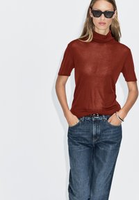 Rollkragen-T-Shirt in Rostfarbe aus leichtem Stoff, mit kurzen Ärmeln und legerer Passform, kombiniert mit dunkelblauen Jeanshosen.