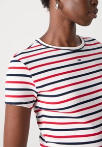T-shirt rayé en blanc, avec des rayures horizontales rouges et bleu marine, texture côtelée et encolure ronde. Comprend un petit logo.