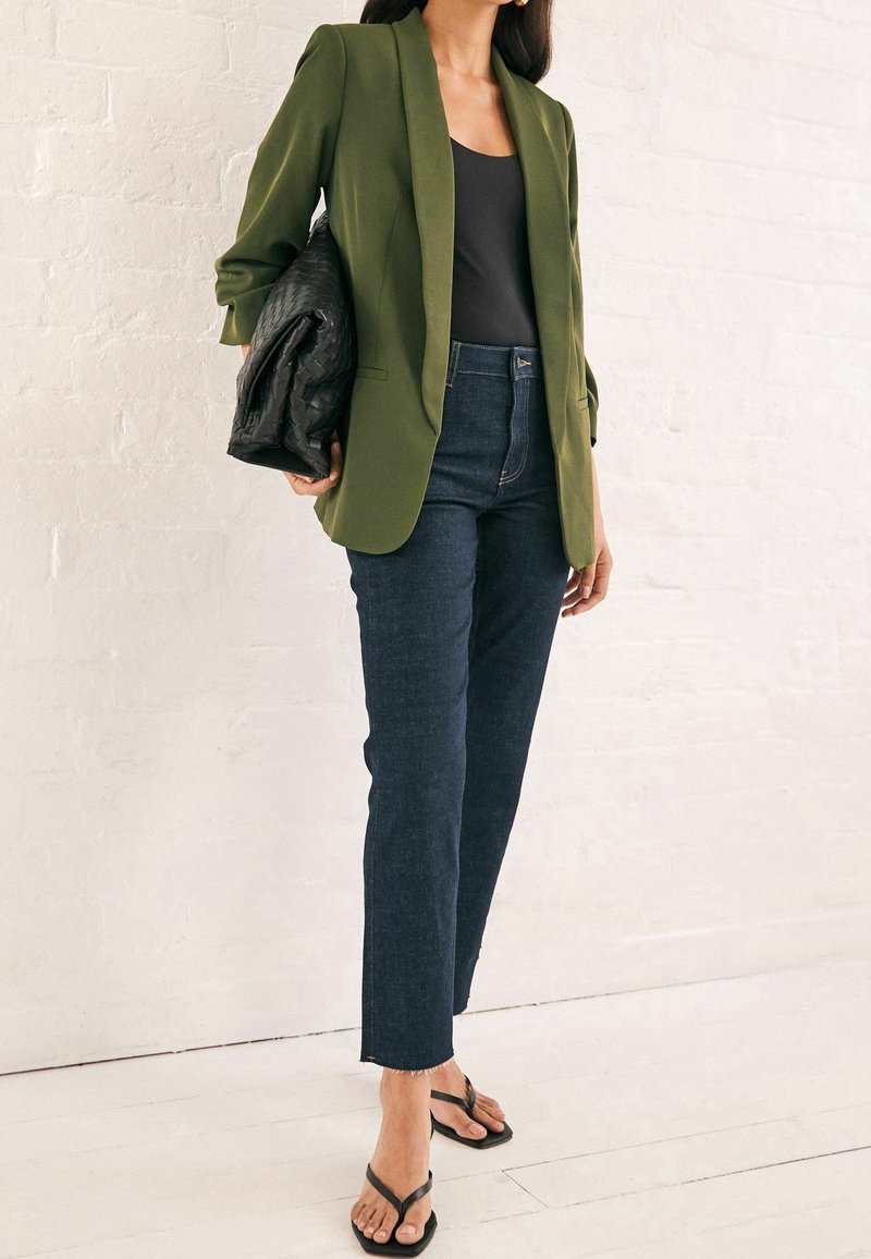 Femme portant un blazer vert olive, un débardeur noir, un jean bleu foncé, des tongs noires, et tenant une grande pochette tissée noire.
