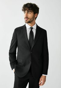 Man met krullend haar en baard, gekleed in een zwart pak, wit overhemd en zwarte stropdas, staand met één hand in de zak, kijkt zijwaarts.