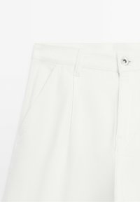 Pantalon blanc montrant la zone de la taille avec passants de ceinture, poche latérale et fermeture à bouton argenté sur un fond uni.