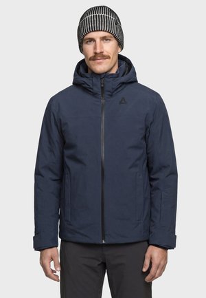 Mann trägt eine marineblaue Winterjacke mit Kapuze und eine schwarz-weiß gestreifte Strickmütze, steht vor einem schlichten Hintergrund.
