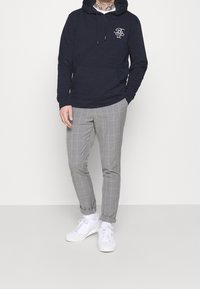 Muž v námořnické mikině s logem, šedých kostkovaných kalhotách, bílých ponožkách a bílých teniskách, stojící na neutrálním světlém pozadí.