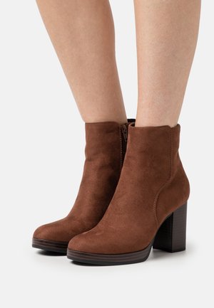 Bottes chevilles en suède marron avec un bout rond, une fermeture éclair latérale et un talon bloc épais. Texture lisse et détails de coutures minimalistes.