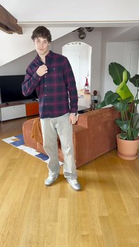 Rot und marineblau karierter Hemd mit Knöpfen, hellgraue Jeans, silberne Sneaker. Die Szene umfasst eine Topfpflanze und ein strukturiertes braunes Sofa.