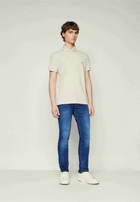 Young man standing, wearing a beige polo shirt, blue slim-fit jeans, and white sneakers against a plain light background.