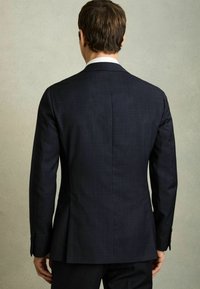 Tumesinine ülikonnajakk tekstuurse kangaga, slim fit disain, üks taga vent, ja kahe nupuga kinnitus; koos valge ülikonnakaelusega särgiga.