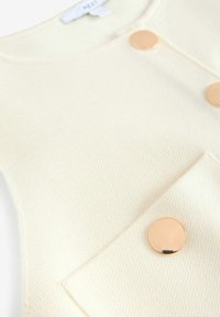 Gilet di colore crema con colletto rotondo, dotato di due bottoni dorati e particolare tasca, che evidenzia un design pulito e moderno.