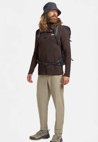 Zwarte jas met ritssluiting en een aansluitend ontwerp, kaki broek, grijze wandelschoenen en een zwarte bucket hat. Uitgerust met een rugzak voorzien van bandjes.