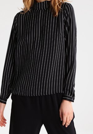 Woman wearing a black long-sleeve blouse with white vertical stripes and black pants against a plain background.