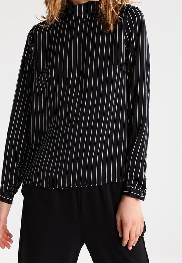 Femme portant un chemisier noir à manches longues avec des rayures verticales blanches et un pantalon noir sur un fond uni.