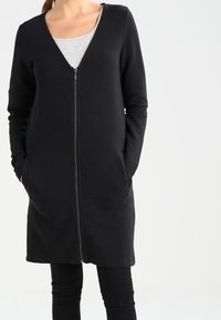 Cardigan noir à manches longues avec un col en V et une fermeture éclair frontale intégrale, doté de poches latérales et d'un tissu lisse et texturé.