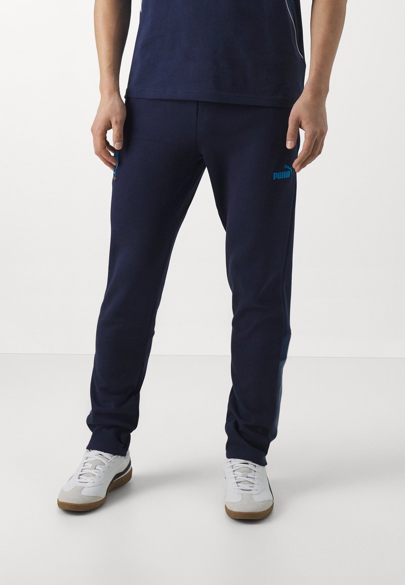 Navy blue sweatpants made of soft fabric, featuring a slim design, with a turquoise Puma logo on the left leg and side pockets.
