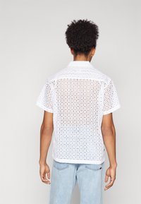 White short-sleeve shirt with a grid-like open pattern, collared design, and relaxed fit, paired with light blue denim jeans.