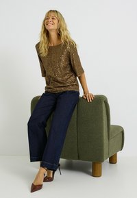 Top de lentejuelas bronce con escote redondo y mangas tres cuartos, combinado con pantalones de mezclilla oscuros de pierna ancha y zapatos de tacón burdeos.