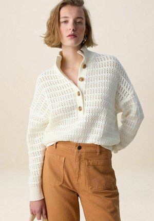 Jeune femme portant un pull en maille blanc avec des boutons et un pantalon marron taille haute, debout devant un fond neutre.