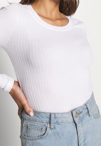 Bílý žebrovaný top s dlouhým rukávem a kulatým výstřihem, kombinovaný se světle modrými džíny s vysokým pasem, které mají šité kapsy a zapínání na knoflík.