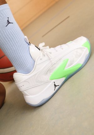 Witte basketbalschoen met groene accenten, gedragen met lichtblauwe Jordan-sokken op een binnenbaan naast een basketbal.