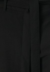 Mustan värinen kangashihna, sileä tekstuuri, kiinnitetty vyötäröltä korkeisiin housuihin, joissa on etuseam ja puhdas, minimalistinen muotoilu.