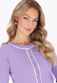 Lila cardigan med en smal passform, rund halsringning, kontrasterande vit kant och knappknäppning i guldfärg längs placket. Mjuk stickad textur.