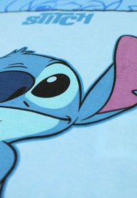 Tissu en coton bleu avec le visage d'un personnage de dessin animé, aux grands yeux expressifs et accents roses sur les oreilles. Le texte indique "STITCH".