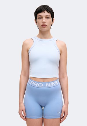 Giovane persona che indossa un crop top senza maniche azzurro chiaro e pantaloncini aderenti abbinati con fascia "NIKE PRO", in piedi davanti a uno sfondo chiaro e uniforme.