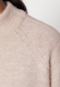 Ljust rosa stickad tröja med hög krage, texturerad garn och en lätt ludden yta. Raglanärmar med en avslappnad passform.