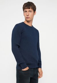 Pull en tricot bleu marine avec une texture côtelée, col rond et manches longues, associé à un jean foncé. Le design présente une coupe classique.