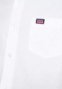 Chemise blanche à boutons avec une poche poitrine comportant un écusson logo GANT en bleu marine, blanc et rouge.
