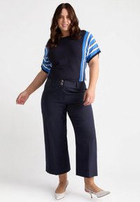 Granatowe spodnie w stylu dzwonów z podwójnym guzikiem z przodu, w połączeniu z granatową bluzką z niebieskimi i białymi paskami na krótkich rękawach.
